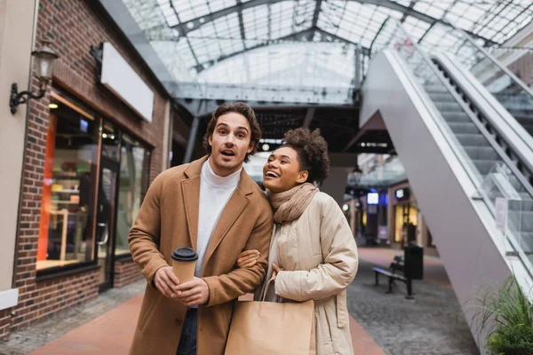 Amazed multiethnic couple with purchases walking in mall — Stock Photo