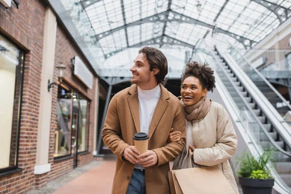 Cheerful multiethnic couple with purchases walking in shopping mall — Stock Photo