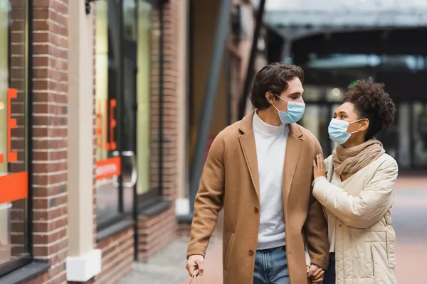 Multiethnic couple in medical masks holding hands while walking in shopping mall — Stock Photo