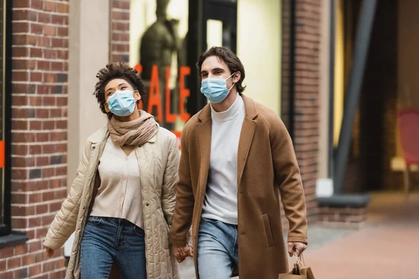 Joven pareja interracial en máscaras médicas cogidas de la mano mientras camina en el centro comercial - foto de stock