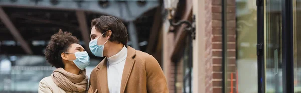 Giovane coppia interrazziale in maschere mediche guardando l'un l'altro nel centro commerciale, banner — Foto stock