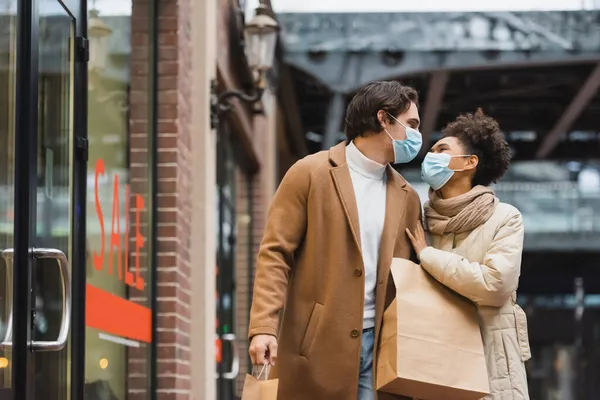 Jovem casal interracial em máscaras médicas olhando uns para os outros e andando com sacos de compras no shopping — Fotografia de Stock