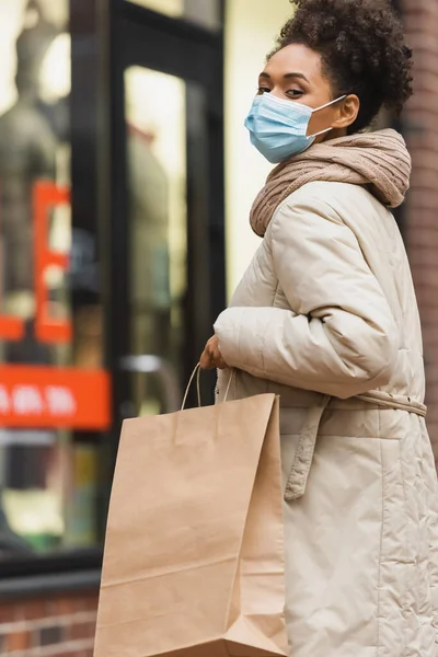 Брюнетка африканская американка в медицинской маске держит сумку с покупками — стоковое фото
