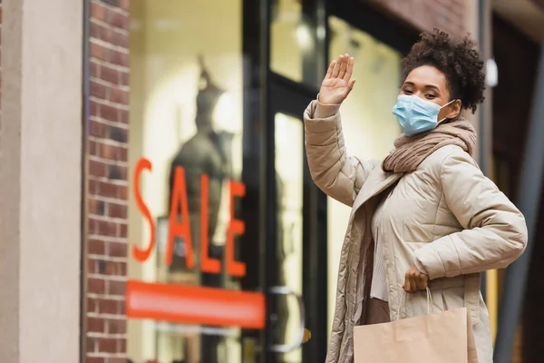 Morena afroamericana americana mujer en máscara médica sosteniendo bolsa de compras y saludando de la mano en el centro comercial - foto de stock