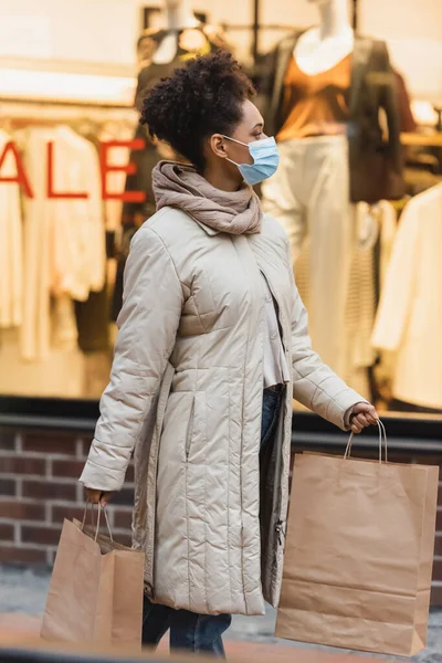 Morena afroamericana americana mujer en máscara médica caminando con bolsas de compras en el centro comercial - foto de stock