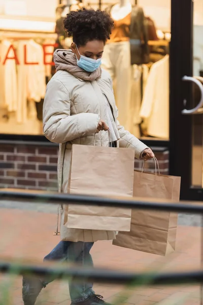 Morena afro-americana em máscara médica segurando sacos de compras no shopping — Fotografia de Stock