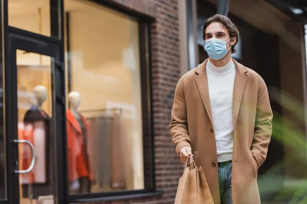 Hombre joven en máscara médica y abrigo beige caminando con compras en el centro comercial - foto de stock