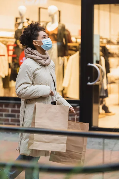 Joven afroamericana mujer en máscara médica caminando con compras en el centro comercial - foto de stock
