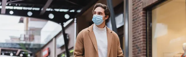 Giovane in maschera medica e cappotto beige passeggiando nel centro commerciale, banner — Foto stock