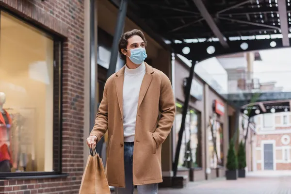 Jeune homme en masque médical et manteau beige marchant avec des sacs à provisions dans le centre commercial — Photo de stock