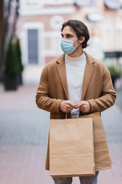 Jovem em máscara médica e casaco bege segurando compra no shopping center — Fotografia de Stock