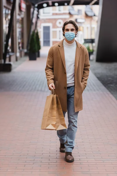 Mann mit medizinischer Maske und Mantel läuft mit Einkaufstüten in Einkaufszentrum — Stockfoto