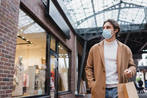 Jeune homme en masque médical et manteau beige marchant avec sac à provisions dans le centre commercial — Photo de stock