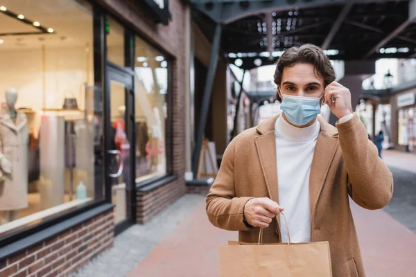 Jeune homme ajustant masque médical et tenant sac à provisions dans le centre commercial — Photo de stock