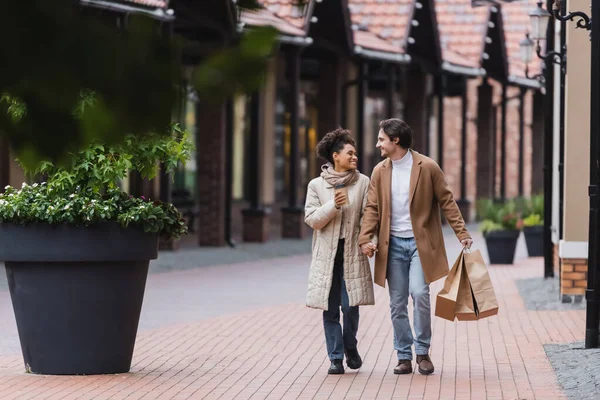 Happy multiethnic couple in coats holding hands while walking with purchases near mall — Stock Photo