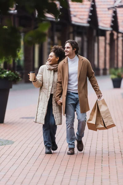 Alegre pareja multiétnica en abrigos tomados de la mano mientras camina con compras cerca del centro comercial - foto de stock