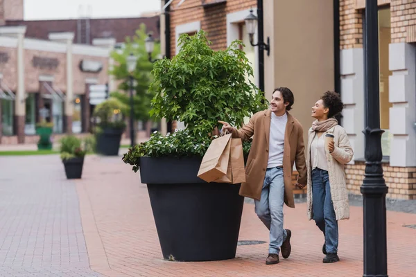 Cheerful man pointing with finger and holding hands with african american woman while walking with purchases near mall — Stock Photo
