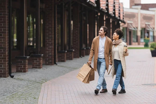 Pareja multiétnica positiva en abrigos tomados de la mano mientras camina con compras cerca del centro comercial - foto de stock