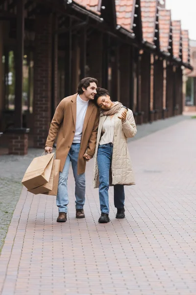 Любящая многонациональная пара в пальто, держащаяся за руки во время прогулки с покупками возле торгового центра — стоковое фото