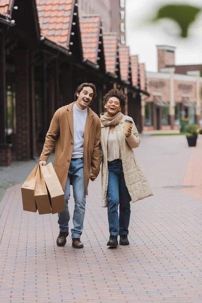 Excitada pareja multiétnica en abrigos tomados de la mano mientras camina con compras cerca del centro comercial - foto de stock