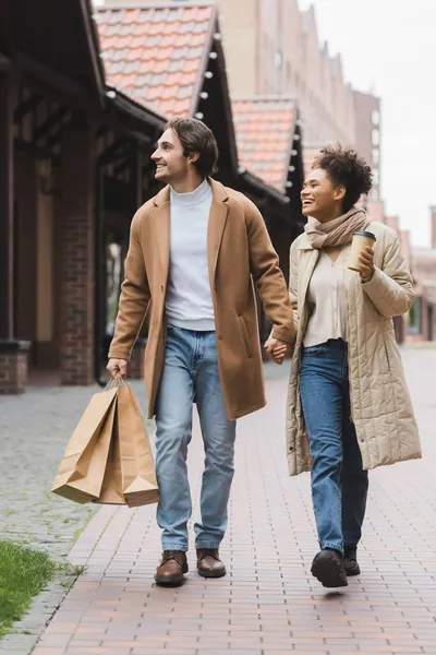 Joyful multiethnic couple in coats holding hands while walking with purchases near mall — Stock Photo