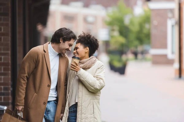 Fröhliche afrikanisch-amerikanische Frau hält Pappbecher neben lächelnden Freund mit Einkaufstüten — Stockfoto