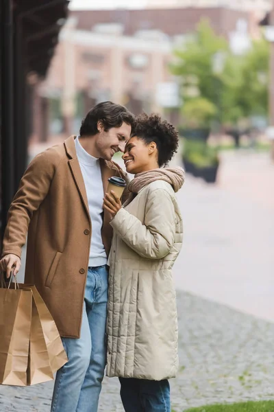 Pleased african american woman holding paper cup near smiling boyfriend with shopping bags — Stock Photo