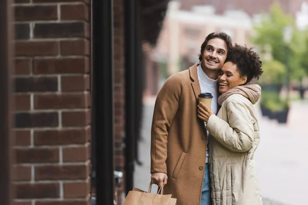 Freudige afrikanisch-amerikanische Frau hält Pappbecher neben glücklichem Freund mit Einkaufstaschen — Stockfoto