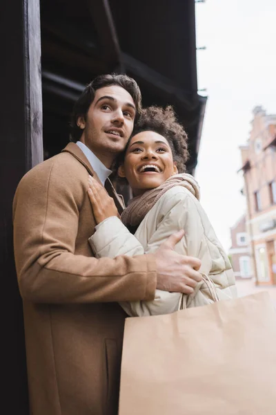 Vista de ángulo bajo de la mujer afroamericana emocionada abrazando novio con bolsas de compras - foto de stock