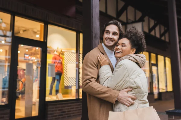 Feliz africana americana mujer abrazando alegre novio con bolsas de compras - foto de stock