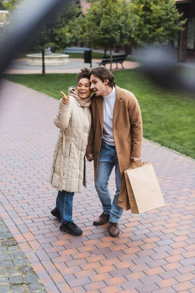 Happy african american woman pointing away near cheerful boyfriend with shopping bags outside — Stock Photo