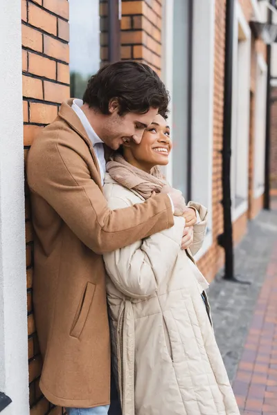 Glücklicher Mann umarmt fröhliche afrikanisch-amerikanische Freundin in der Nähe von Gebäude im Freien — Stockfoto