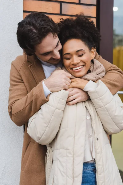 Alegre hombre abrazando alegre africano americano novia cerca de la construcción al aire libre — Stock Photo