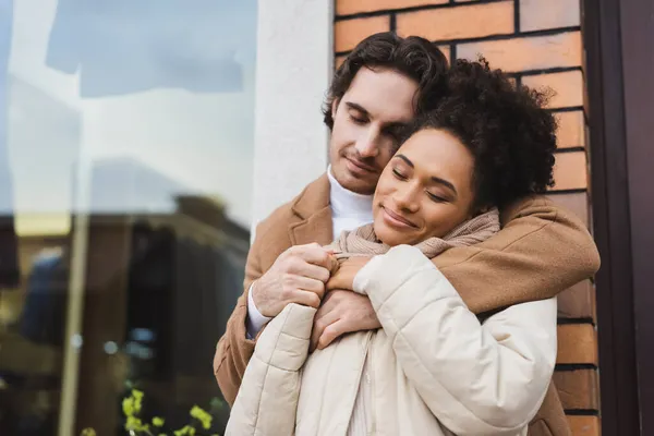 Zufriedener Mann umarmt fröhliche afrikanisch-amerikanische Freundin in der Nähe eines Gebäudes im Freien — Stockfoto