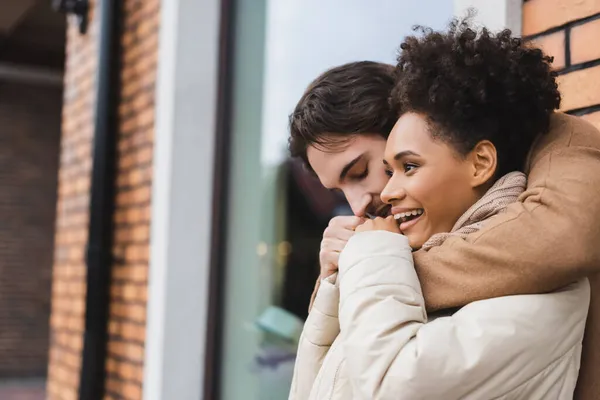 Young man embracing and warming up cheerful african american girlfriend near building outdoors — Stock Photo