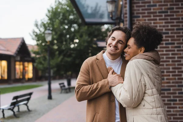 Positiver Mann umarmt fröhliche afrikanisch-amerikanische Freundin in der Nähe von Gebäude im Freien — Stockfoto