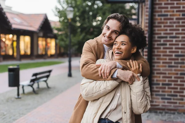 Glücklicher Mann umarmt aufgeregte afrikanisch-amerikanische Freundin in der Nähe eines Gebäudes im Freien — Stockfoto