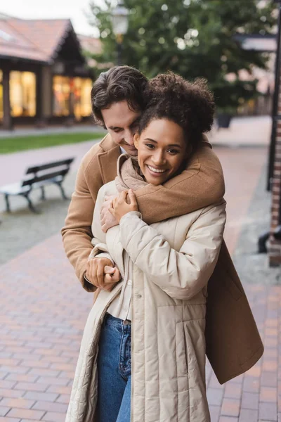 Smiling man embracing happy african american girlfriend near building outdoors — Stock Photo