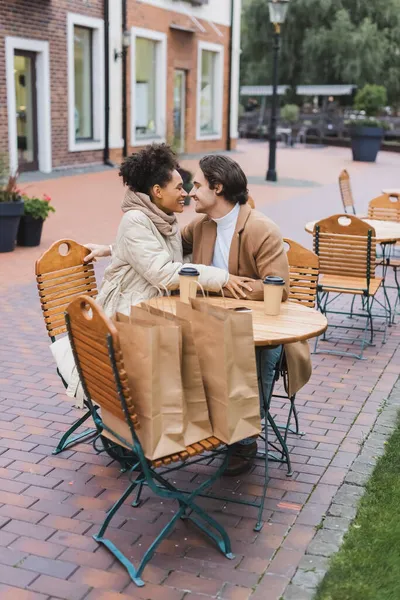 Happy interracial couple sitting near paper cups and shopping bags in outdoor cafe — Stock Photo