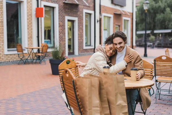 Счастливая межрасовая пара обнимается возле бумажных стаканчиков и сумок для покупок в кафе на открытом воздухе — стоковое фото