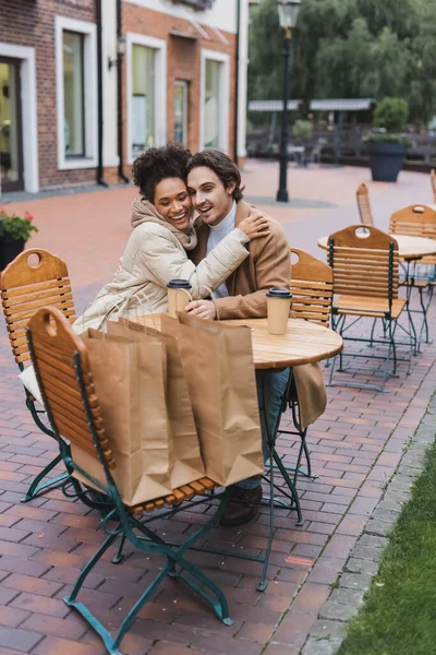 Feliz casal interracial abraçando perto de copos de papel e sacos de compras no café ao ar livre — Fotografia de Stock