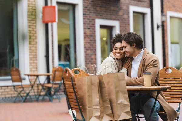 Cheerful interracial couple embracing near paper cup and shopping bags in outdoor cafe — Stock Photo