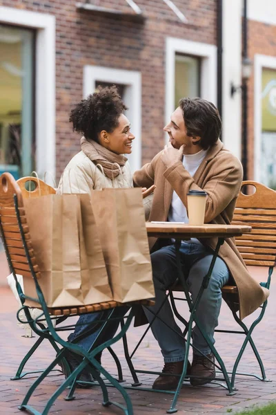 Cheerful interracial couple talking and looking at each other near paper cup and shopping bags in outdoor cafe terrace — Stock Photo