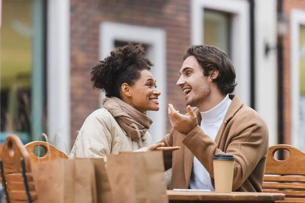 Joyful interracial couple talking and looking at each other near paper cup and shopping bags in outdoor cafe terrace — Stock Photo