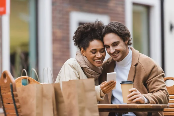 Mujer afroamericana feliz sosteniendo teléfono inteligente cerca de novio con taza de papel - foto de stock