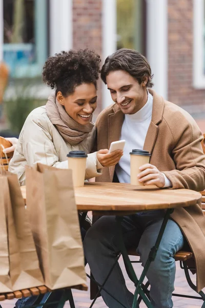 Compiaciuta donna afro-americana che tiene smartphone vicino al fidanzato con tazza di carta — Foto stock