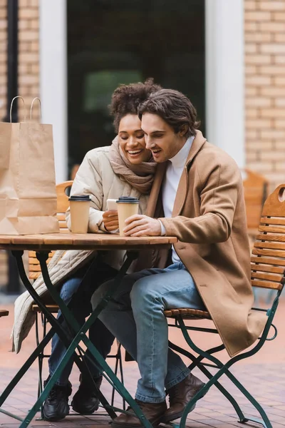 Compiaciuta donna afroamericana che tiene smartphone vicino al fidanzato con tazza di carta e shopping bag sul tavolo — Foto stock