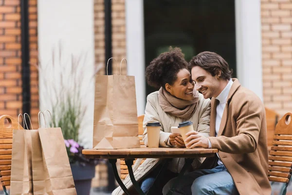 Pleased african american woman holding smartphone and looking at boyfriend with paper cup — Stock Photo