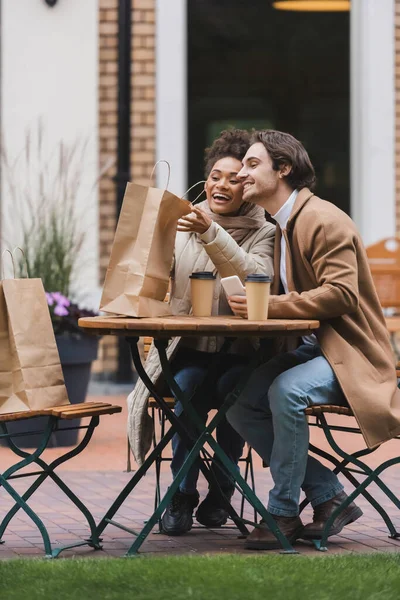 Mujer afroamericana feliz sosteniendo bolsa de compras cerca de novio con teléfono inteligente - foto de stock