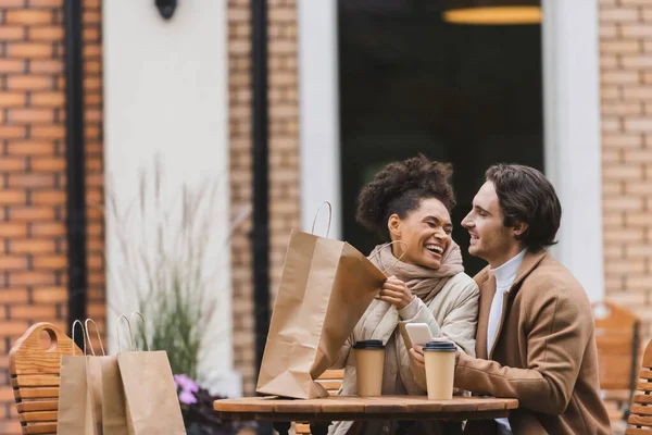 Счастливая африканская американка держит сумку с покупками и смеется рядом с парнем со смартфоном — стоковое фото